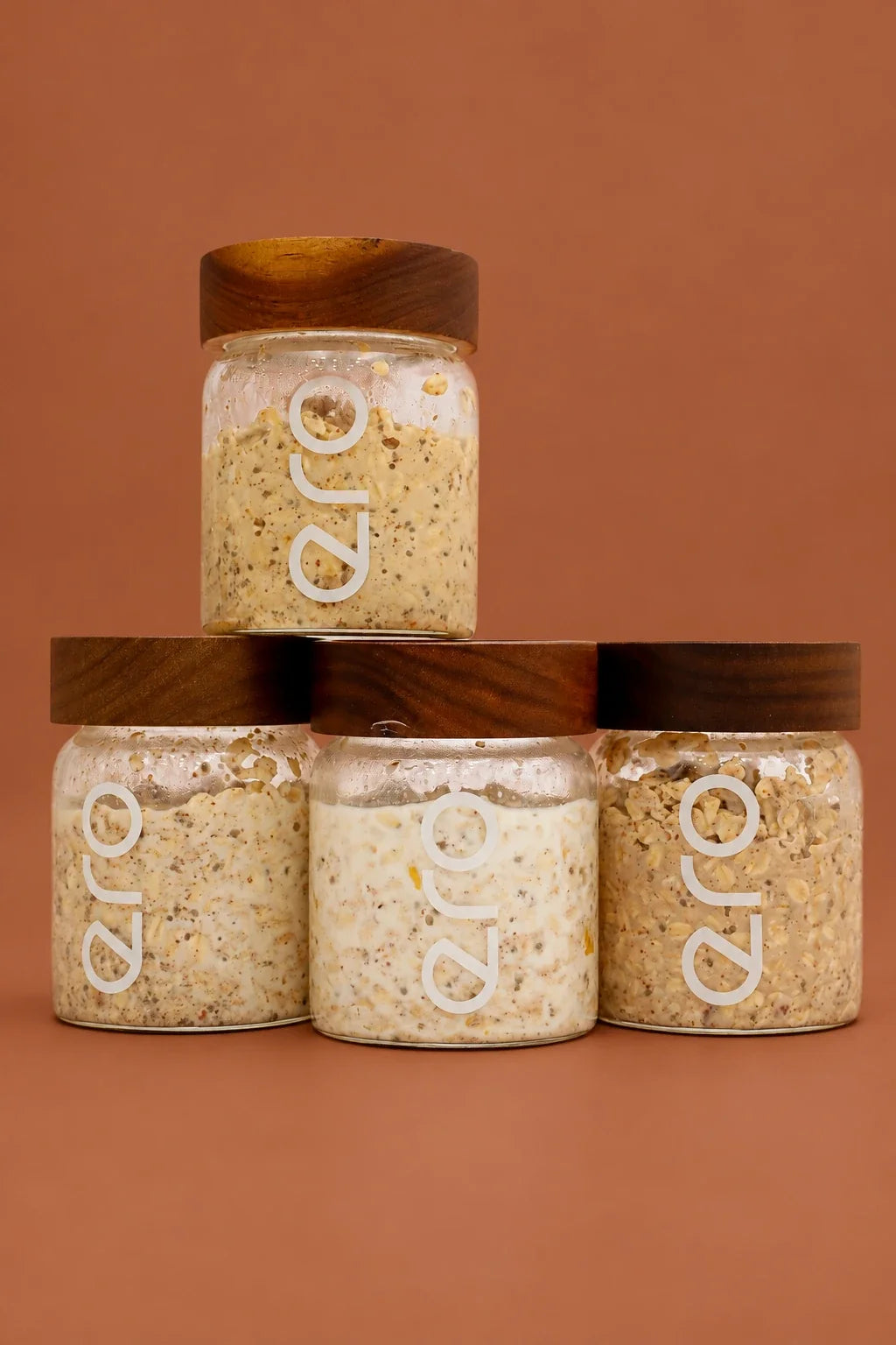Four glass jars of prepared ero overnight oats with wooden lids, stacked on a rust background