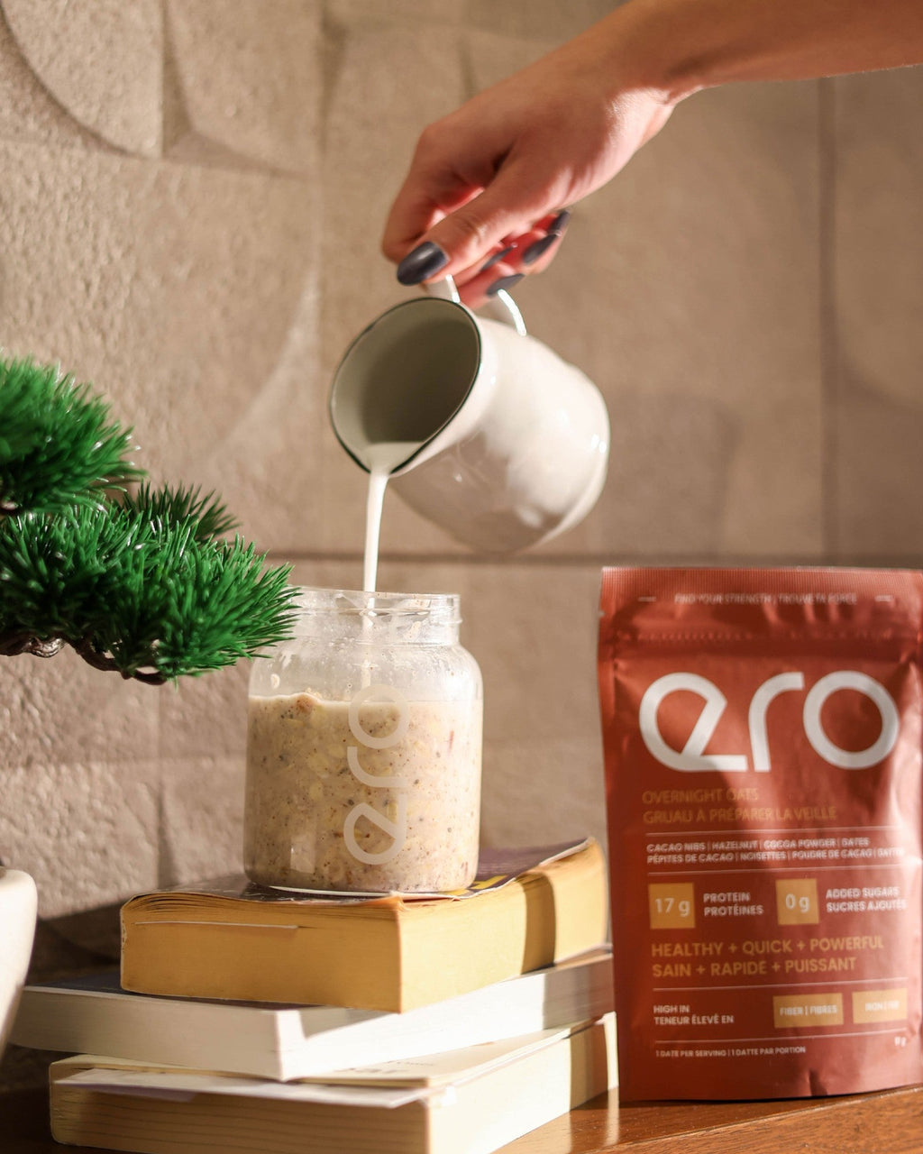 Pouring milk into a glass jar of ero Cacao, Hazelnut & Dates overnight oats, with the pouch beside it on a wooden shelf