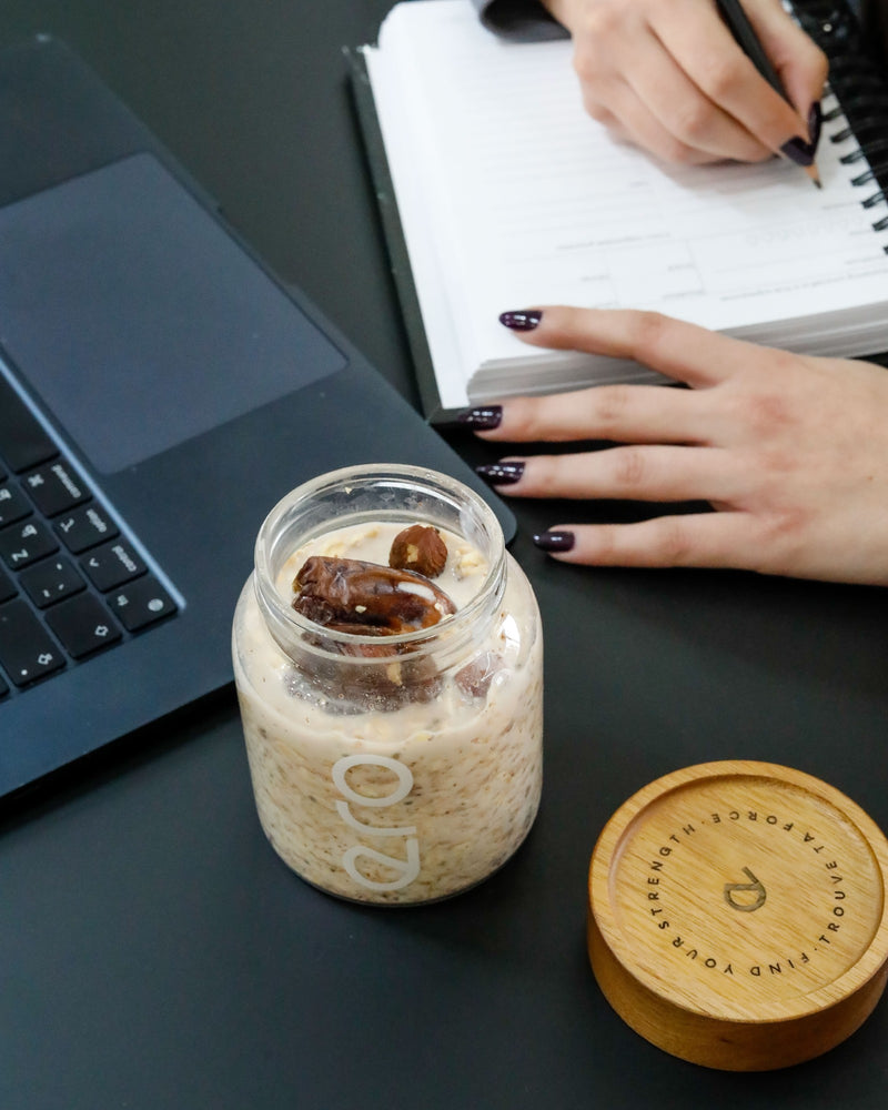 ero overnight oats in a glass jar with dates and hazelnuts, on a desk beside a laptop and planner