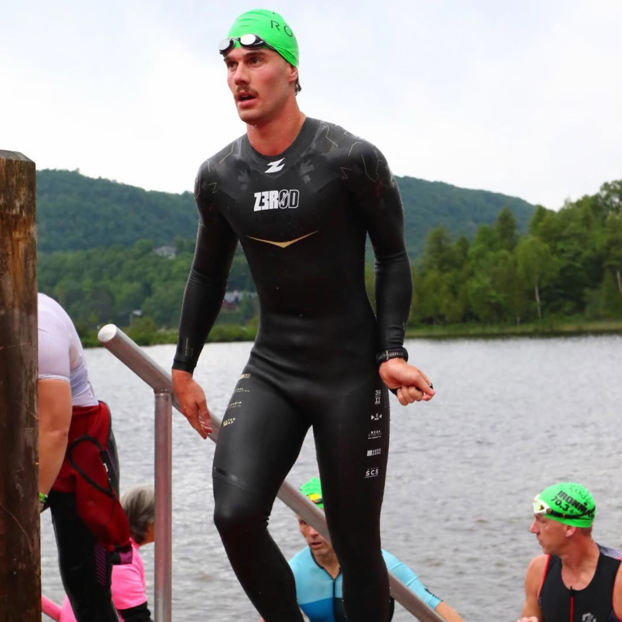 Man in a black wetsuit with a green swim cap standing by a lake, representing open water swimming, triathlon training, and active lifestyle for Ero Evergreen supplements.